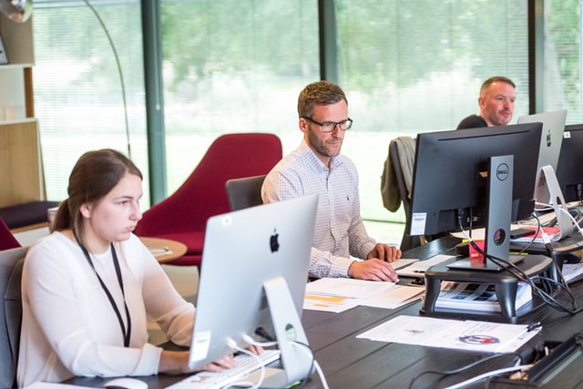 Two men and one woman sitting in front of computers at work with large windows in the background.