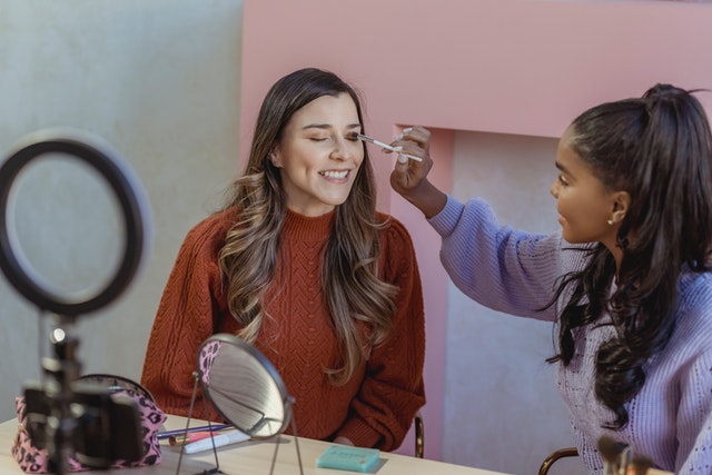 Two women record themselves sitting behind a magnifying glass while one puts eyeshadow on another