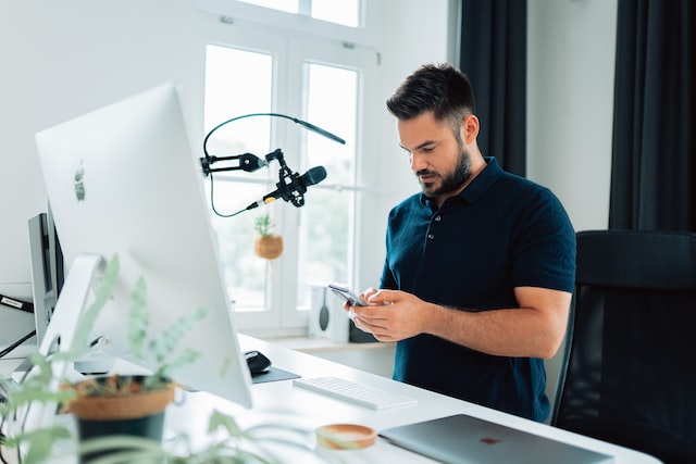 Man typing on smartphone in front of large monitor and podcast mic