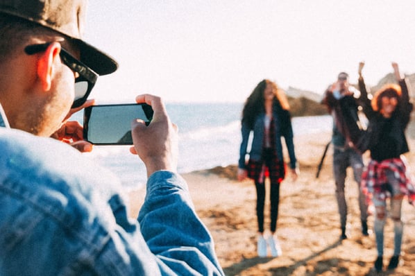 Man in blue shirt and hat taken picture of friends on the beach with mobile phone.