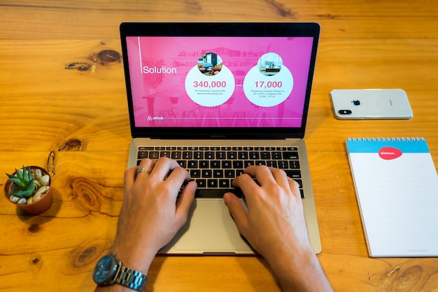 Two hands typing on a computer with pink textured background and white concentric circles with statistics against wooden table with other flat lay photo objects.