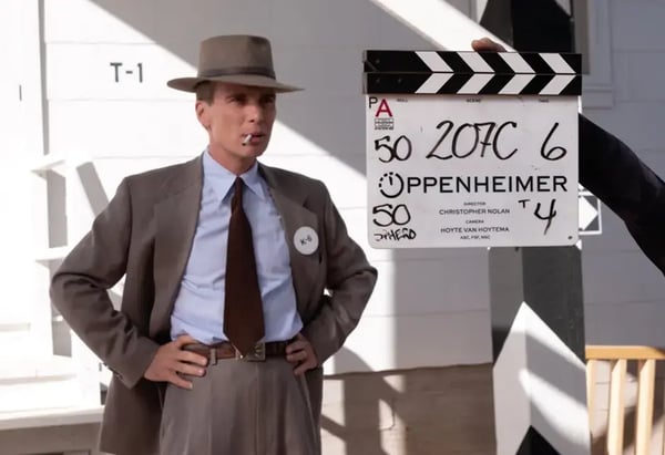 Man in a brown suit and fedora posing with a cigarette in his mouth with a clapperboard showcasing the start of the scene.