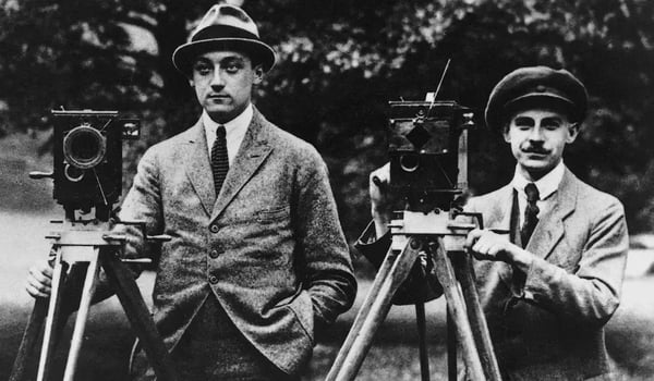 Black-and-white image of two men dressed in formal wear alongside cameras shooting film.