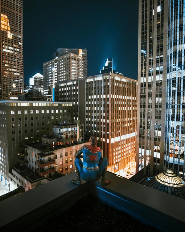 Spider-Man crouching on a roof ledge of a high-rise building at night with the skyline lit up.