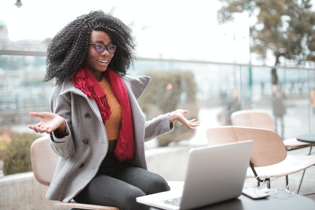 Woman with orange sweater, red scarf, and gray coat smiling into laptop video with hands out in happy gesture